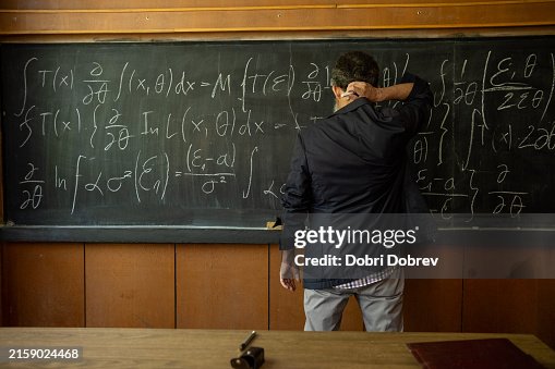 A teacher with a long white beard, big gray hair, and glasses is wearing a checkered shirt and sports jacket in front of a blackboard with complex formulas written on it. A teacher with a long white beard, big gray hair, and glasses is wearing a checkered shirt and sports jacket in front of a blackboard with complex formulas written on it.