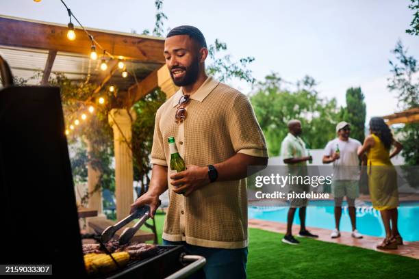 hombre asando comida mientras sostiene una cerveza en una fiesta de barbacoa al aire libre junto a la piscina. grupo de amigos socializando en el fondo. concepto de celebración de verano. plano medio - a la parrilla fotografías e imágenes de stock
