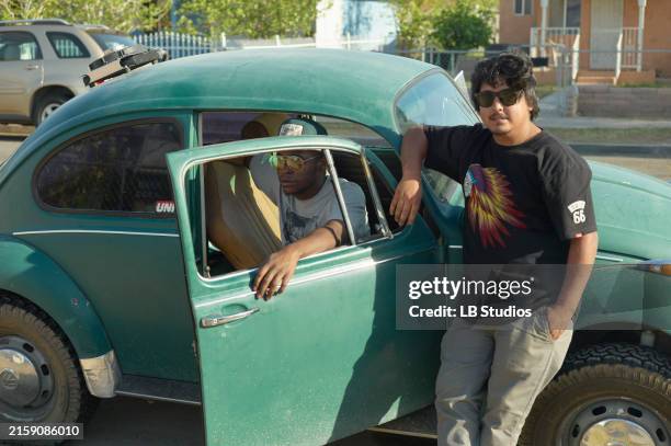 two young men are standing next to a vintage green volkswagen beetle, one sitting inside with the door open and the other leaning on the door from outside - vintage car stock pictures, royalty-free photos & images