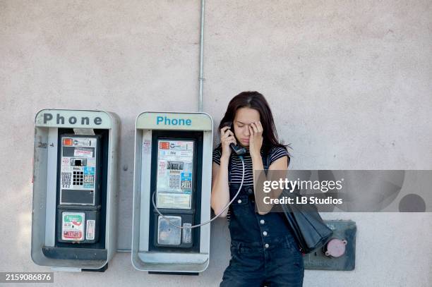 a woman is standing between two public payphones, engaged in a conversation on one of them, with her eyes closed and a slight frown, possibly indicating a serious or troubling discussion - pay phone stock pictures, royalty-free photos & images
