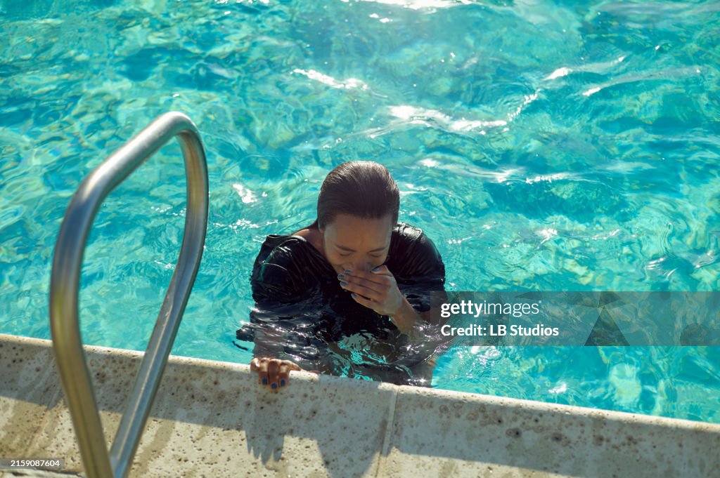 A person with wet hair is immersed in a swimming pool, holding onto the edge near a pool ladder, blowing bubbles in the water
