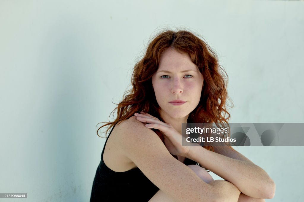 A red-haired woman with a subtle gaze sits against a white wall, her head resting on her hand