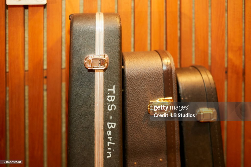 Two upright guitar cases are standing against a wooden-slatted background, one green with a white stripe and the other brown, both featuring shiny clasps