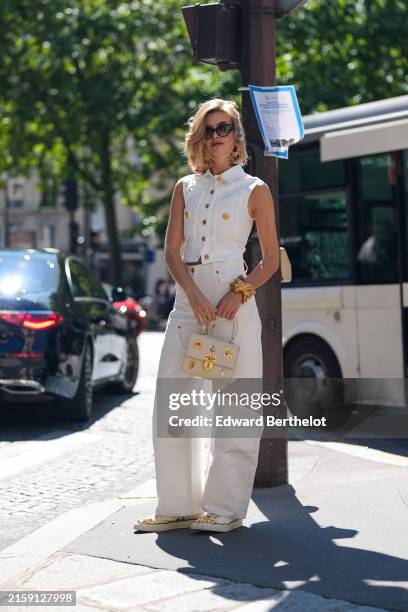 Xenia Adonts wears black sunglasses, gold Schiaparelli earrings, white and gold button up tailored vest, gold bracelet, white loose denim jean pants,...