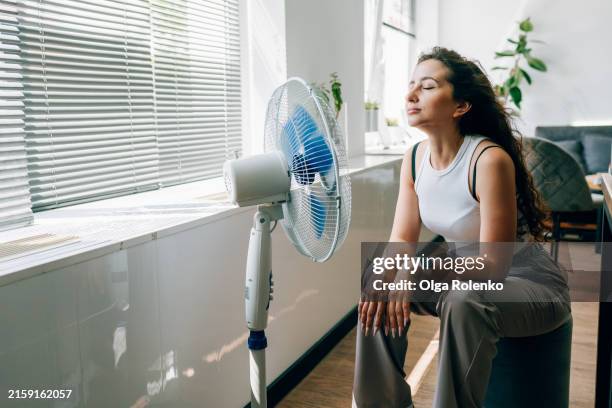 cooling off: long haired woman sits in front of fan to beat the heat at home - hittegolf stockfoto's en -beelden