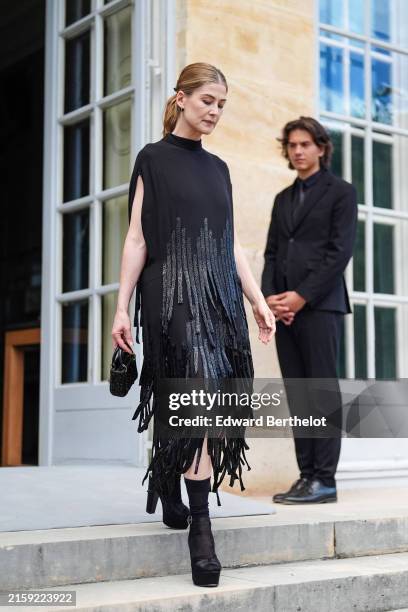 Rosamund Pike wears black earrings, black and shiny black pattern tasseled sleeveless midi Dior dress, shiny black sequin Dior bag, black socks,...