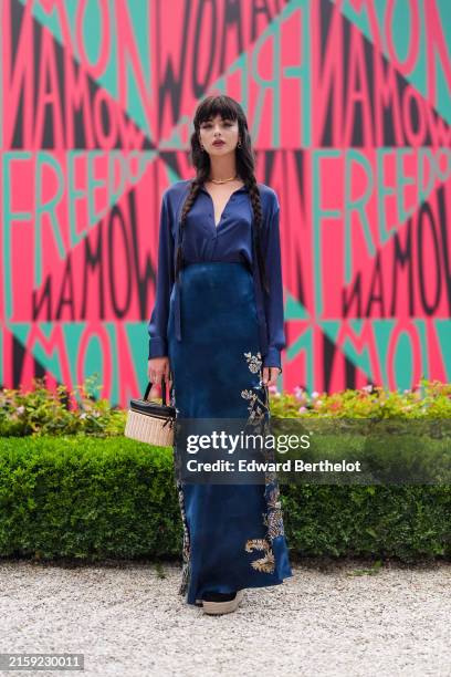 Deva Cassel wears silver earrings, gold necklace, dark blue satin button up long sleeve half unbuttoned, navy blue maxi skirt with silver and gold...