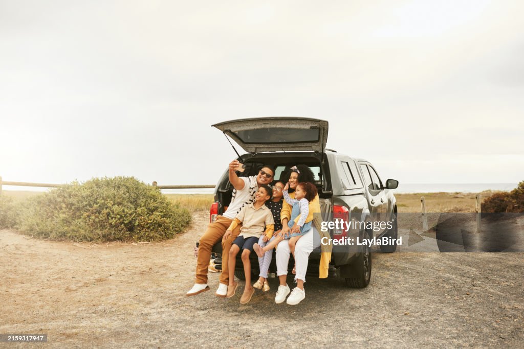 Des parents heureux avec des enfants prenant un selfie avec un téléphone portable à l’arrière du véhicule à la plage