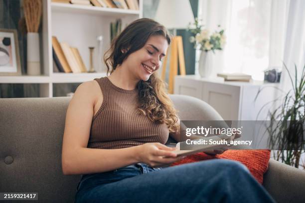 donna sorridente che legge un romanzo su un divano accogliente - libro in brossura foto e immagini stock