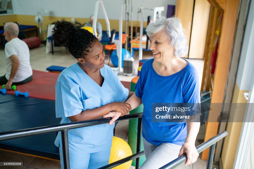Senior woman guided by therapist in walking exercise between bars at rehab
