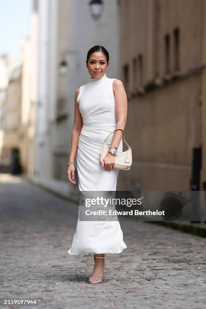 Izabel Sim, stylist, wears silver earrings, white draped sleeveless midi dress, silver bracelets, a watch, beige leather bag, shiny silver heels with...