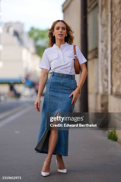 Claudia Sulewski wears gold earrings, white half button up short sleeve Patou shirt, dark brown Patou leather bag, a gold bracelet, washed denim jean...