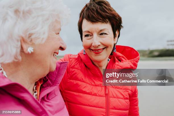 a woman puts her arm around her elderly mother as they enjoy a walk on a windy beach - encouragement stock pictures, royalty-free photos & images