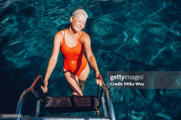 woman with short white hair in a bright orange swimsuit climbing a ladder out of clear blue water, smiling. - badkläder bildbanksfoton och bilder