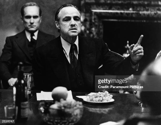 American actor Marlon Brando gestures at a table while American actor Robert Duvall sits behind him in a still from the film, 'The Godfather,'...