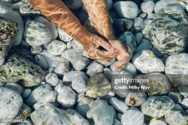 hands in shallow water casting shadows over smooth pebbles, creating a beautiful underwater scene. - vadear fotografías e imágenes de stock