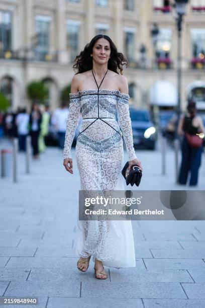 Guest wears white off the shoulder long sleeve sheer mesh lace pattern maxi Giambattista Valli dress, shiny black leather bag with gold chain, gold...