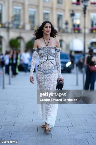 Guest wears white off the shoulder long sleeve sheer mesh lace pattern maxi Giambattista Valli dress, shiny black leather bag with gold chain, gold...