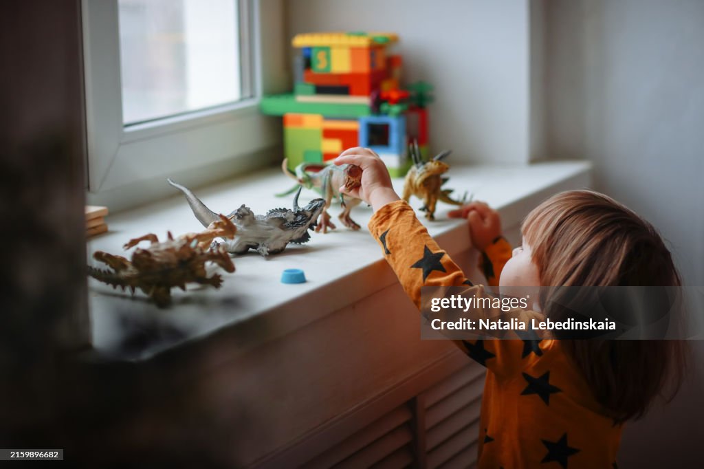 Mysterious Playtime: Toddler Enjoying Dinosaur Toys on Windowsill, Surrounded by a Dark and Cozy Home Environment