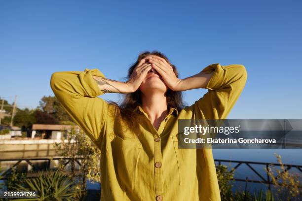 a woman in a green shirt covering her face with her hands outdoors. background includes greenery and a blue sky. - hands covering eyes stock pictures, royalty-free photos & images