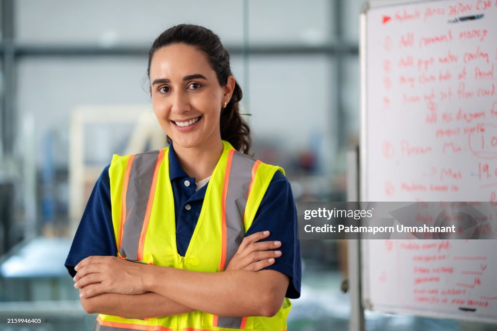 A determined robotics engineer, wearing reflective clothing, stands in an industrial factory with arms crossed, staring intently into the camera. Professional's confidence and expertise, highlighting the challenges and dedication involved in role.