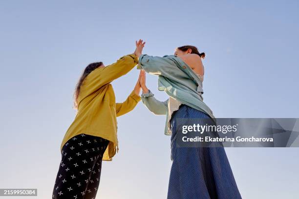 two women high-fiving outdoors against a clear blue sky. - vida sencilla fotografías e imágenes de stock