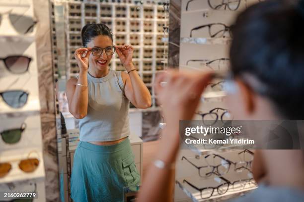beautiful woman choosing glasses in optical store - instrumento óptico fotografías e imágenes de stock