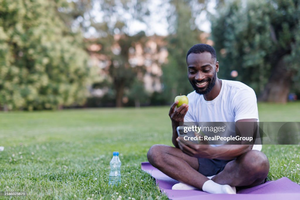 Man enjoying apple and phone on yoga mat outside