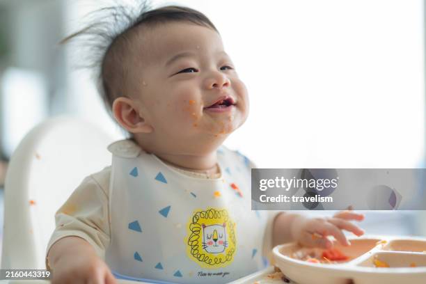 adorable asian baby boy eating food from the plate by himself while sitting in high chair cropped hand of mother feeding her baby boy with spoon in high chair at home baby milestone, growth and development concept - baby bib stock pictures, royalty-free photos & images