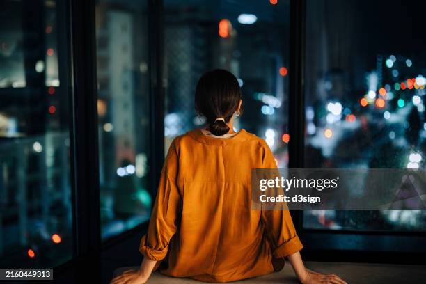 rear view of young asian woman relaxing at cozy home, sitting alone in the living room looking over through window the spectacular illuminated city skyline at night - insomnia stock-fotos und bilder