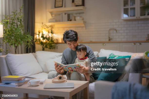 night family lifestyle the adorable little baby boy focuses on the book while his dad is teach him a book story at night asian father and his son read children's book together on sofa in living room home interior background - storytelling stock pictures, royalty-free photos & images