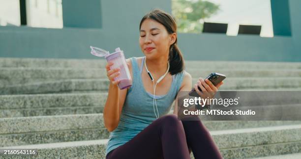 young asian female athlete runner drinking protein shake and using smartphone for fitness motivation after exercise workout in urban park. diet and healthy food workout exercise. - drink milkshake stock pictures, royalty-free photos & images