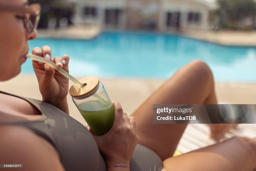 Eine nicht wiederzuerkennende Touristin trinkt Detox mit einem Trinkhalm aus einem Glas und sonnt sich am Swimmingpool