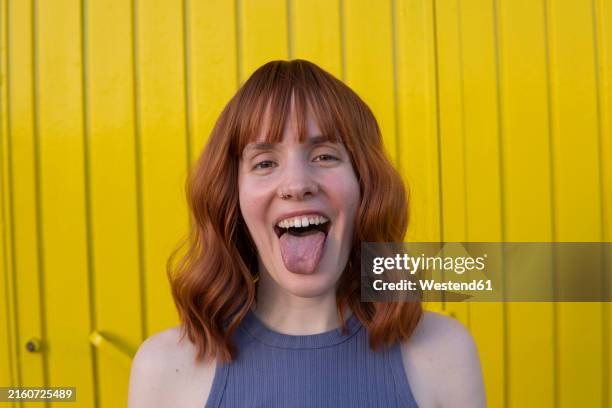 happy redhead woman sticking tongue out in front of yellow wall - mensentong stockfoto's en -beelden