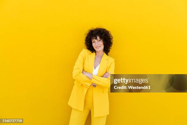 smiling beautiful woman wearing blazer standing with arms crossed in front of yellow wall - yellow suit stock pictures, royalty-free photos & images