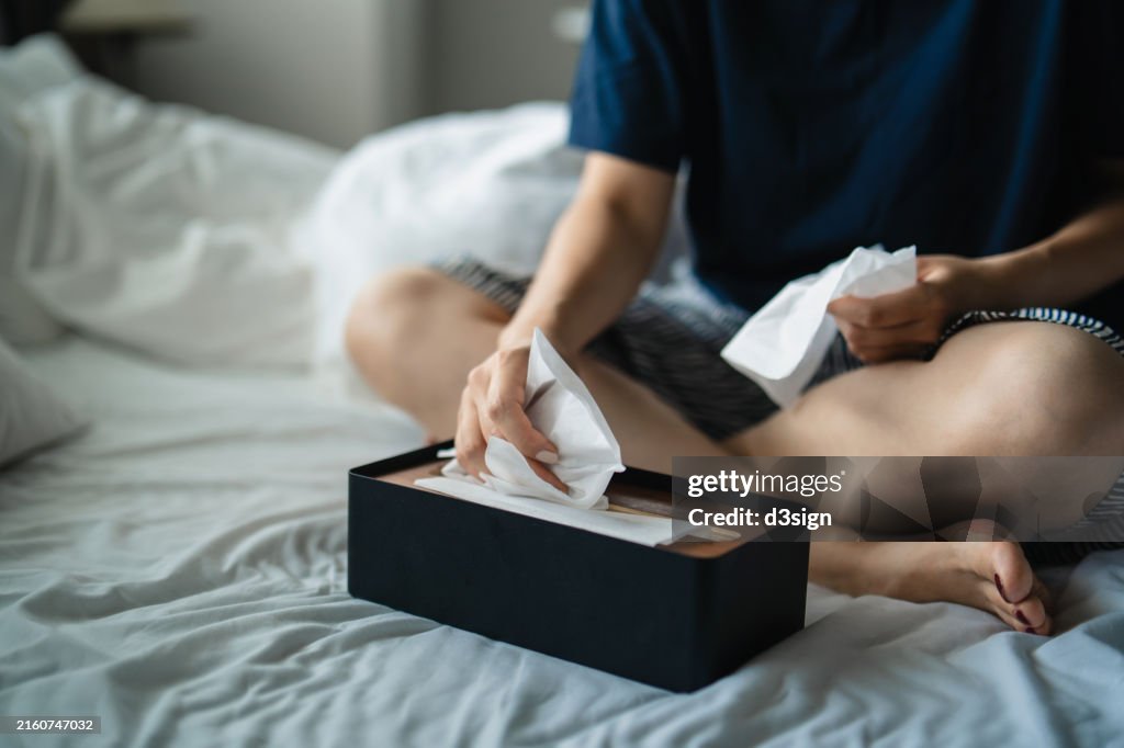 Cropped shot of woman feeling ill, suffering from cold, flu and allergy, grabbing facial tissues from the box on bed. Concept of health and illness
