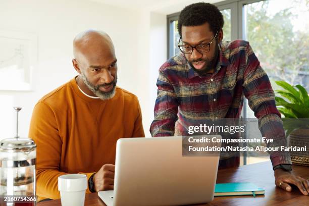 young man and mature man using laptop in kitchen - hot desking stock pictures, royalty-free photos & images