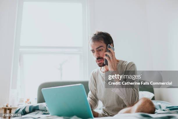 a man using a laptop and smart phone at the same time, sits on a bed, looking confused - fraud stock pictures, royalty-free photos & images