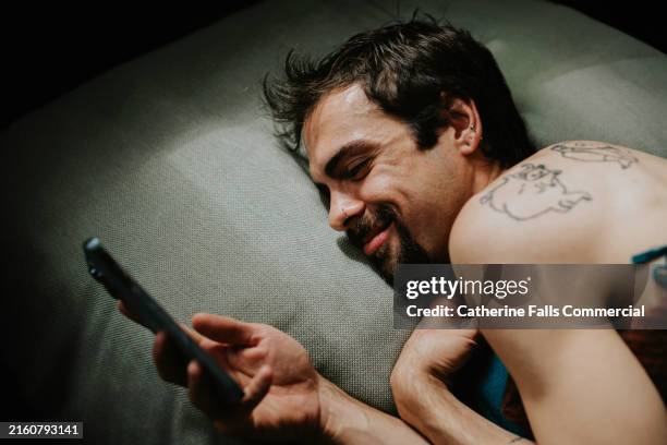a man looks smitten as he giggles whilst looking at his smart phone whilst lying in bed - romantic good night images stock pictures, royalty-free photos & images