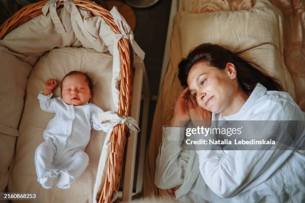 mother relaxing next to newborn in wicker cradle in the bedroom, authentic motherhood moments - straws crib stock pictures, royalty-free photos & images