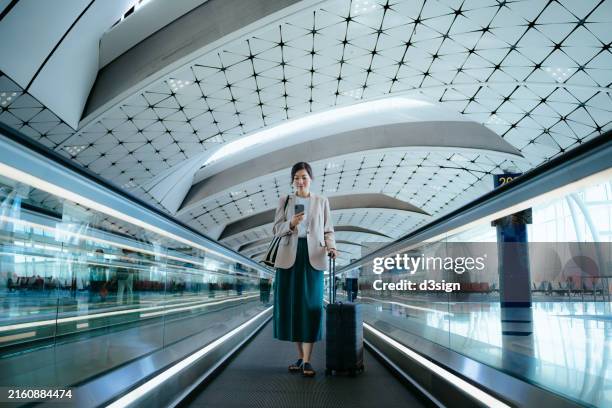 young asian businesswoman with suitcase using smartphone while walking on moving walkway at airport terminal. business traveller. lifestyle and technology. travel and vacation concept - abflugbereich stock-fotos und bilder