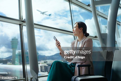 Confident young Asian businesswoman with suitcase looking ahead while using smartphone waiting for her flight at airport terminal. Business traveller. Woman in business. Business lifestyle and technology. Travel and vacation concept Confident young Asian businesswoman with suitcase looking ahead while using smartphone waiting for her flight at airport terminal. Business traveller. Woman in business. Business lifestyle and technology. Travel and vacation concept