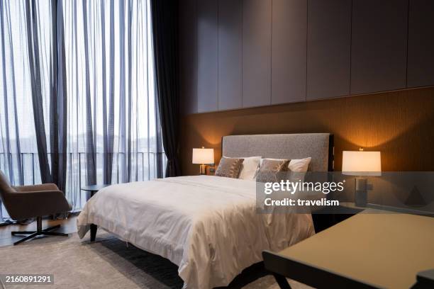 interior of a contemporary cozy bedroom with furniture in earthtone colour scheme a cozy armchair, and a small writing desk, all bathed in natural sunlight filtering through sheer curtains. - hotel stock-fotos und bilder