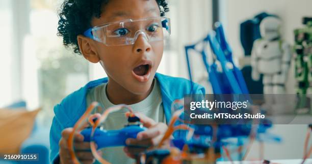 primer plano de un joven afroamericano que usa el control remoto para un proyecto de robótica en medio de varias herramientas y modelos tecnológicos en casa. cursos de robótica en línea de stem education. - robótica fotografías e imágenes de stock