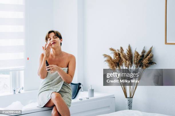 young woman wrapped in a towel applies skincare products on her face while seated by the bathtub. - me time stock pictures, royalty-free photos & images