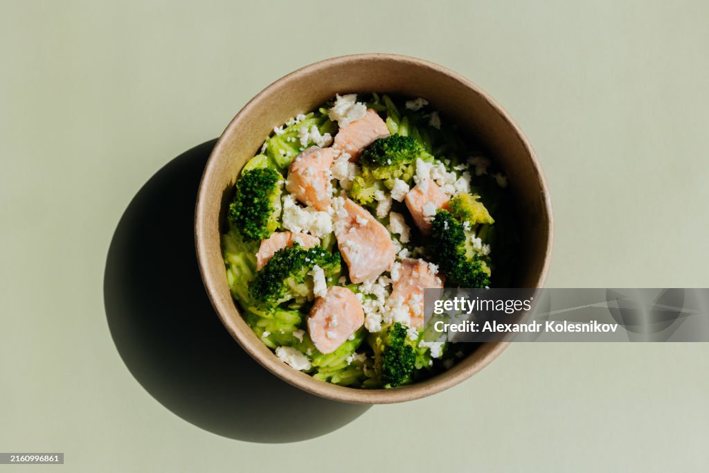 Top view of healthy lunch bowl in reusable box pasta risoni, salmon fillet, broccoli and cheese on green background. Delicious balanced food concept.