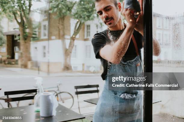 a man wearing an apron, cleans a large window at the front of the cafe where he works - view-through-restaurant-window stock pictures, royalty-free photos & images
