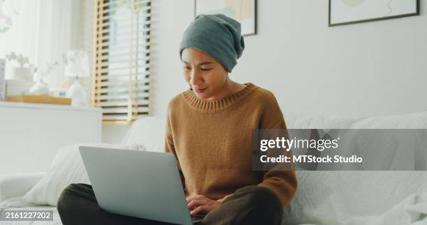 junge krebskranke asiatin mit kopftuch, die bequem an ihrem laptop arbeitet, sitzt auf einem sofa in einem gut beleuchteten, mit pflanzen geschmückten raum. leben mit krebs. - überleben stock-fotos und bilder