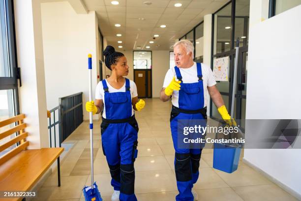 female janitor black ethnicity and senior caucasian male janitor walking through the school hall while carrying cleaning equipment - caretaker stock pictures, royalty-free photos & images