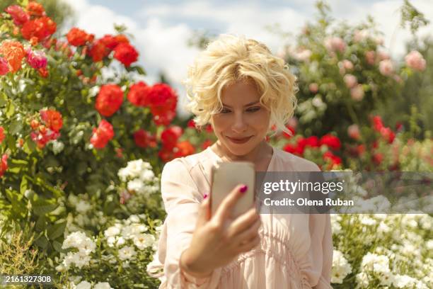 young woman taking selfie among flowers in a summer park. - le telephone rose stock-fotos und bilder